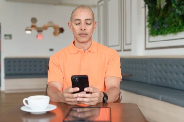 Kafe masasında kahve fincanı ve akıllı telefon kullanan orta yaşlı, turuncu polo tişörtlü yakışıklı kel adam. Teknoloji ve iş hayatı konsepti.