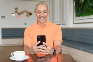 Kafe masasında kahve fincanı ve akıllı telefon kullanan orta yaşlı, turuncu polo tişörtlü yakışıklı kel adam. Teknoloji ve iş hayatı konsepti.