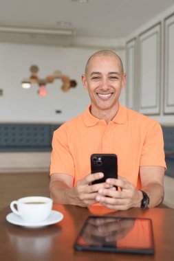 Kafe masasında kahve fincanı ve akıllı telefon kullanan orta yaşlı, turuncu polo tişörtlü yakışıklı kel adam. Teknoloji ve iş hayatı konsepti.