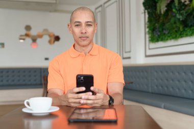 Kafe masasında kahve fincanı ve akıllı telefon kullanan orta yaşlı, turuncu polo tişörtlü yakışıklı kel adam. Teknoloji ve iş hayatı konsepti.