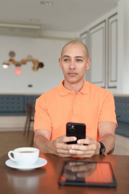Kafe masasında kahve fincanı ve akıllı telefon kullanan orta yaşlı, turuncu polo tişörtlü yakışıklı kel adam. Teknoloji ve iş hayatı konsepti.