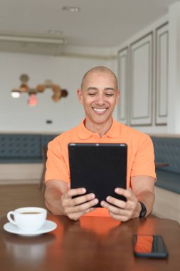 Kafe masasında kahve fincanı takan, kulaklık takan ve dijital tablet bilgisayar kullanan orta yaşlı yakışıklı bir adam. Teknoloji ve günlük yaşam tarzı kavramı.