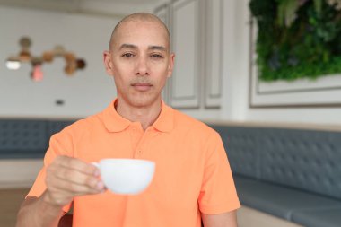 Kafe masasında kahve fincanı tutan orta yaşlı, turuncu polo tişörtlü yakışıklı, kel bir İtalyan. Günlük yaşam tarzı kavramı.