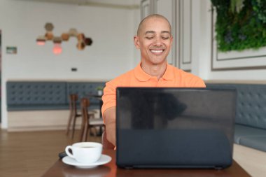 Kafe masasında kahve fincanı ile kapalı mekanda dizüstü bilgisayarla çalışan orta yaşlı, kel, turuncu polo tişörtlü bir adam. Uzak çalışma ve teknoloji yaşam tarzı konsepti.