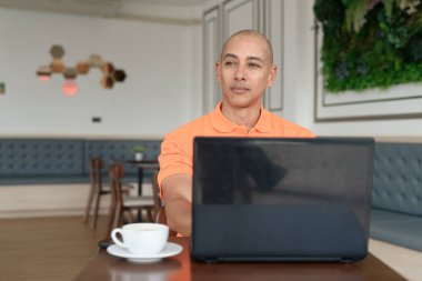 Kafe masasında kahve fincanı ile kapalı mekanda dizüstü bilgisayarla çalışan orta yaşlı, kel, turuncu polo tişörtlü bir adam. Uzak çalışma ve teknoloji yaşam tarzı konsepti.