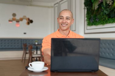 Kafe masasında kahve fincanı ile kapalı mekanda dizüstü bilgisayarla çalışan orta yaşlı, kel, turuncu polo tişörtlü bir adam. Uzak çalışma ve teknoloji yaşam tarzı konsepti.