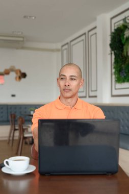 Kafe masasında kahve fincanı ile kapalı mekanda dizüstü bilgisayarla çalışan orta yaşlı, kel, turuncu polo tişörtlü bir adam. Uzak çalışma ve teknoloji yaşam tarzı konsepti.