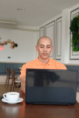 Kafe masasında kahve fincanı ile kapalı mekanda dizüstü bilgisayarla çalışan orta yaşlı, kel, turuncu polo tişörtlü bir adam. Uzak çalışma ve teknoloji yaşam tarzı konsepti.