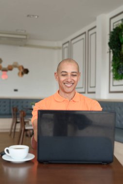 Kafe masasında kahve fincanı ile kapalı mekanda dizüstü bilgisayarla çalışan orta yaşlı, kel, turuncu polo tişörtlü bir adam. Uzak çalışma ve teknoloji yaşam tarzı konsepti.