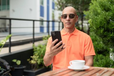 Yakışıklı, orta yaşlı, kel, turuncu polo tişörtlü, açık büfe masasında oturan, güneş gözlüğü takan ve cep telefonu kullanan bir adam. Günlük yaşam tarzı kavramı.