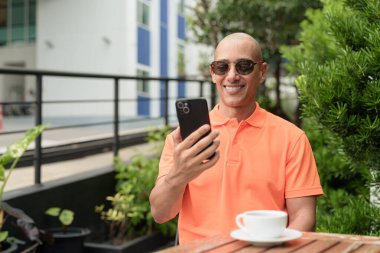 Yakışıklı, orta yaşlı, kel, turuncu polo tişörtlü, açık büfe masasında oturan, güneş gözlüğü takan ve cep telefonu kullanan bir adam. Günlük yaşam tarzı kavramı.