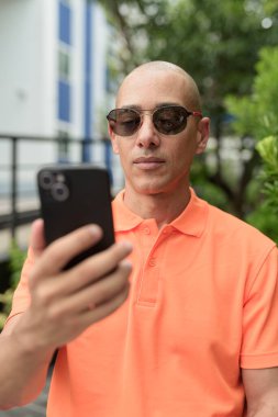 Yakışıklı, orta yaşlı, kel, turuncu polo tişörtlü, açık büfe masasında oturan, güneş gözlüğü takan ve cep telefonu kullanan bir adam. Günlük yaşam tarzı kavramı.