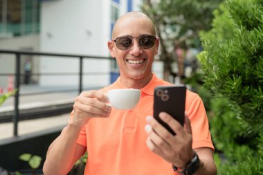 Yakışıklı, orta yaşlı, kel, turuncu polo tişörtlü, açık büfe masasında oturan, güneş gözlüğü takan ve cep telefonu kullanan bir adam. Günlük yaşam tarzı kavramı.