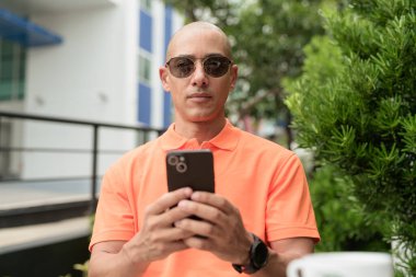 Yakışıklı, orta yaşlı, kel, turuncu polo tişörtlü, açık büfe masasında oturan, güneş gözlüğü takan ve cep telefonu kullanan bir adam. Günlük yaşam tarzı kavramı.