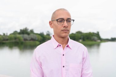 40 'lı yaşlarda, kel, olgun, pembe iş gömleği ve gözlük takan bir erkek doğa parkında gölün kenarında duruyor. Rahatlama, açık hava yaşam tarzı, rahatlık ve refah kavramı..