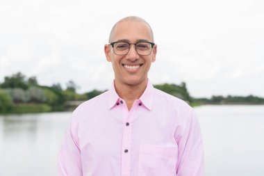 40 'lı yaşlarda, kel, olgun, pembe iş gömleği ve gözlük takan bir erkek doğa parkında gölün kenarında duruyor. Rahatlama, açık hava yaşam tarzı, rahatlık ve refah kavramı..