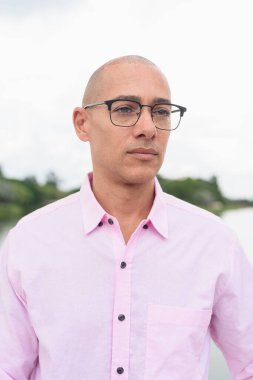 40 'lı yaşlarda, kel, olgun, pembe iş gömleği ve gözlük takan bir erkek doğa parkında gölün kenarında duruyor. Rahatlama, açık hava yaşam tarzı, rahatlık ve refah kavramı..