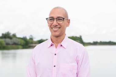 40 'lı yaşlarda, kel, olgun, pembe iş gömleği ve gözlük takan bir erkek doğa parkında gölün kenarında duruyor. Rahatlama, açık hava yaşam tarzı, rahatlık ve refah kavramı..