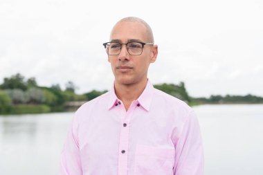 40 'lı yaşlarda, kel, olgun, pembe iş gömleği ve gözlük takan bir erkek doğa parkında gölün kenarında duruyor. Rahatlama, açık hava yaşam tarzı, rahatlık ve refah kavramı..