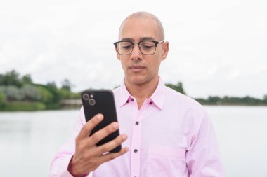 40 'lı yaşlarda, kel, olgun, pembe iş gömleği ve gözlük takan bir erkek doğa parkında gölün kenarında duruyor. Rahatlama, açık hava yaşam tarzı, rahatlık ve refah kavramı..