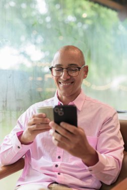 40 'lı yaşlarda kel, yetişkin bir erkek modern kahve dükkanında pembe günlük iş gömleği ve gözlük takıyor şehir yaşam tarzı, eğlence, kafe kültürü ve sosyal rahatlama kavramı..