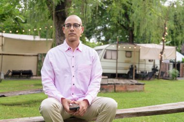 40 'lı yaşlarda kel, olgun bir erkek pembe gömlek ve gözlük takıyor. Kamp alanı çevresi ve karavan geçmişi var. Açık hava eğlence ortamında seyahat, turizm ve yaşam tarzı kavramı.