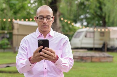 40 'lı yaşlarda kel, olgun bir erkek pembe gömlek ve gözlük takıyor. Kamp alanı çevresi ve karavan geçmişi var. Açık hava eğlence ortamında seyahat, turizm ve yaşam tarzı kavramı.