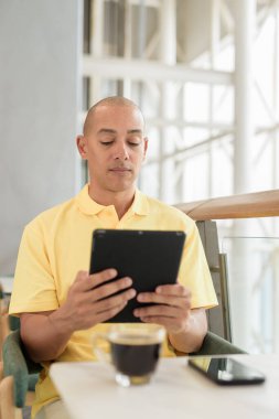 Gündüzleri dijital tablet bilgisayar kullanarak kahve molası verirken şık bir kafede oturan kendinden emin, kel bir İtalyan. Modern yaşam tarzı ve iş konsepti.