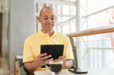 Gündüzleri dijital tablet bilgisayar kullanarak kahve molası verirken şık bir kafede oturan kendinden emin, kel bir İtalyan. Modern yaşam tarzı ve iş konsepti.