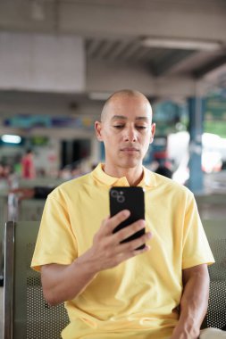 Açık hava otobüs durağında oturan, akıllı telefonuna göz gezdirirken ulaşımı bekleyen olgun, kel bir İspanyol. Tayland 'da seyahat, seyahat ve toplu taşıma kavramı.