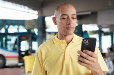 Açık hava otobüs durağında oturan, akıllı telefonuna göz gezdirirken ulaşımı bekleyen olgun, kel bir İspanyol. Tayland 'da seyahat, seyahat ve toplu taşıma kavramı.