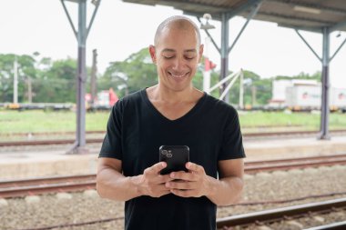 Siyah tişörtlü, olgun, kel bir İspanyol turistin portresi tren istasyonunda telefonla Tayland 'da tren beklerken çekilmiş. Turizm, seyahat ve ulaşım teması.