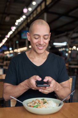 Orta yaşlı, kel bir İspanyol turistin portresi. Açık hava restoranında Tayland eriştesinin tadını çıkarıyor. Yemek yerken gülümsüyor ve yerel mutfağı telefonla deneyimliyor..