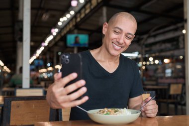 Orta yaşlı, kel bir İspanyol turistin portresi. Açık hava restoranında Tayland eriştesinin tadını çıkarıyor. Yemek yerken gülümsüyor ve yerel mutfağı telefonla deneyimliyor..