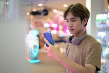 Kahverengi tişörtlü, kulaklıklı Taylandlı genç bir adam. Parlak bir alışveriş merkezinde alışveriş makinesi kullanıyor. Modern yaşam tarzı, teknoloji ve eğlence konsepti..