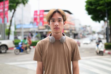 Kulaklıklı yakışıklı Asyalı genç Taylandlı yaz boyunca şehirde modern hayatın tadını çıkarıyor ve kameraya bakıyor.