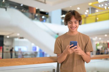 20 yaşında boynunda kulaklıkla Taylandlı bir adam alışveriş merkezinin koridorunda telefonuna gülümserken, mesajlaşırken, online alışveriş yaparken ve şehir yaşamında.