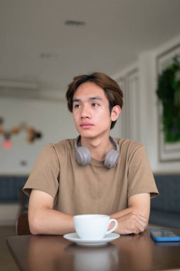 Kahverengi tişörtlü genç Taylandlı öğrenci sosyal medya, uzaktan iletişim ve dijital yaşam tarzı gibi modern bir kafede boynuna kulaklık takıyor..