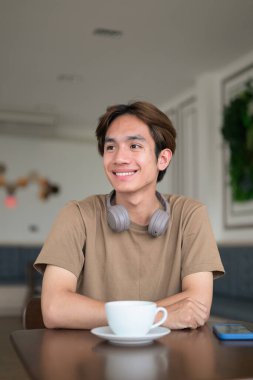 Kahverengi tişörtlü genç Taylandlı öğrenci sosyal medya, uzaktan iletişim ve dijital yaşam tarzı gibi modern bir kafede boynuna kulaklık takıyor..