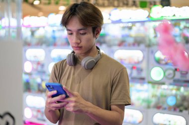 Kahverengi tişörtlü, kulaklıklı Taylandlı genç bir adam. Parlak bir alışveriş merkezinde alışveriş makinesi kullanıyor. Modern yaşam tarzı, teknoloji ve eğlence konsepti..