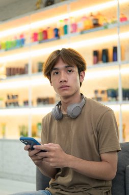 Kahverengi tişörtlü genç Taylandlı öğrenci sosyal medya, uzaktan iletişim ve dijital yaşam tarzı gibi modern bir kafede boynuna kulaklık takıyor..