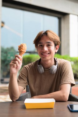 Fast food, aperatif ve yemek konsepti için açık bir masada kızarmış tavuk budu yemeye hazır kulaklıklı, kahverengi tişörtlü genç Asyalı öğrenci.