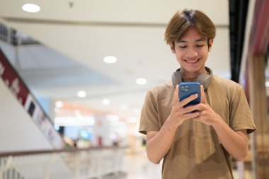 20 yaşında boynunda kulaklıkla Taylandlı bir adam alışveriş merkezinin koridorunda telefonuna gülümserken, mesajlaşırken, online alışveriş yaparken ve şehir yaşamında.