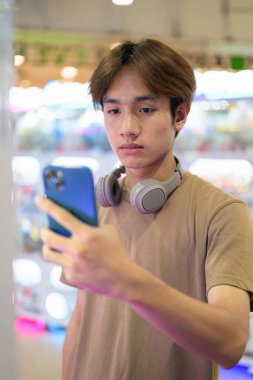 Kahverengi tişörtlü, kulaklıklı Taylandlı genç bir adam. Parlak bir alışveriş merkezinde alışveriş makinesi kullanıyor. Modern yaşam tarzı, teknoloji ve eğlence konsepti..