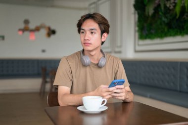 Kahverengi tişörtlü genç Taylandlı öğrenci sosyal medya, uzaktan iletişim ve dijital yaşam tarzı gibi modern bir kafede boynuna kulaklık takıyor..