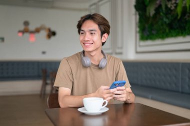 Kahverengi tişörtlü genç Taylandlı öğrenci sosyal medya, uzaktan iletişim ve dijital yaşam tarzı gibi modern bir kafede boynuna kulaklık takıyor..