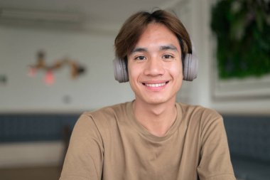 Kahverengi tişörtlü genç Taylandlı öğrenci sosyal medya, uzaktan iletişim ve dijital yaşam tarzı gibi modern bir kafede boynuna kulaklık takıyor..