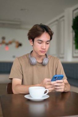 Kahverengi tişörtlü genç Taylandlı öğrenci sosyal medya, uzaktan iletişim ve dijital yaşam tarzı gibi modern bir kafede boynuna kulaklık takıyor..