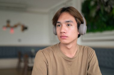 Kahverengi tişörtlü genç Taylandlı öğrenci sosyal medya, uzaktan iletişim ve dijital yaşam tarzı gibi modern bir kafede boynuna kulaklık takıyor..