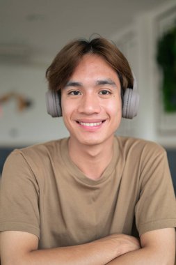 Kahverengi tişörtlü genç Taylandlı öğrenci sosyal medya, uzaktan iletişim ve dijital yaşam tarzı gibi modern bir kafede boynuna kulaklık takıyor..